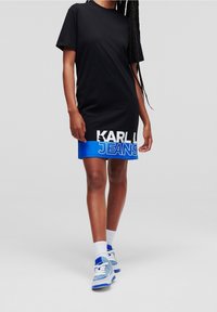 Černé krátké šaty z tričkoviny s nápisem "KARL JEANS" v bílém a modrém tisku na lemu. Spárováno se světle modrými teniskami a bílými ponožkami.