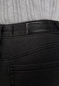 Black denim jeans close-up, featuring a leather label with "HOLLISTER" branding. Ribbed gray knit sweater visible in background.