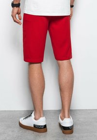 Shorts en coton rouge, longueur genou, avec une texture lisse. Associés à des baskets blanches avec une semelle en caoutchouc marron et des détails en noir.