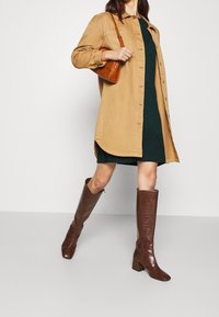 Chemise-jas brown oversize portée sur une robe tricotée vert foncé, associée à des bottes crocodile marron jusqu'aux genoux et à un sac à main marron.