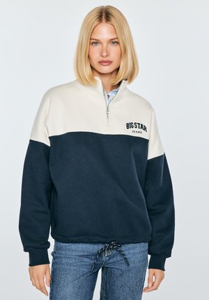 Młoda kobieta z blond bobem, ubrana w biały i granatowy sweter z suwakiem do ćwiartki oraz niebieskie dżinsy, stoi na tle jednolitego tła.