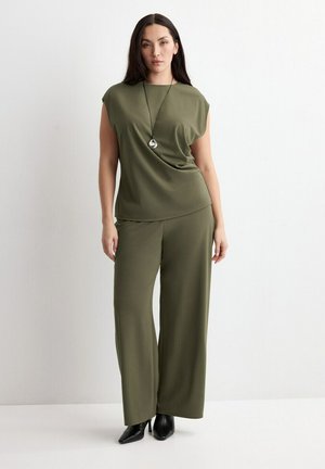 Kvinde iført olivengrøn ærmeløs top og bukser med vide ben, med sorte spidse støvler og sølvvedhæng mod en hvid væg.