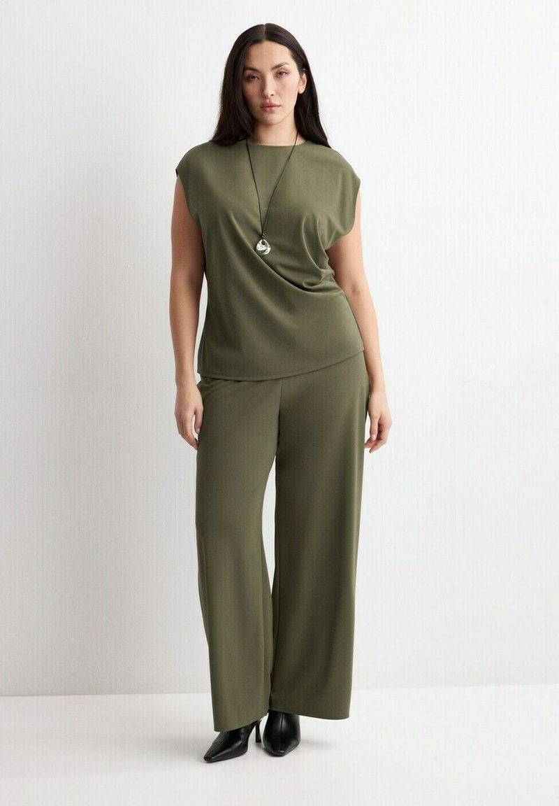 Žena v olivovo zelenom tielku bez rukávov a širokých nohaviciach s čiernymi špicatými čižmami a strieborným náhrdelníkom s príveskom stojí pred bielou stenou.