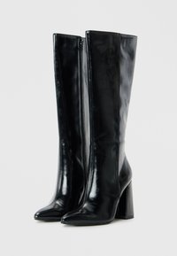Botas de cuero negras brillantes hasta la rodilla, con punta afilada, tacones cuadrados y cremalleras laterales sobre un fondo claro y sencillo.