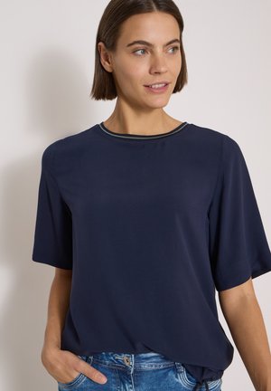Donna con capelli corti e castani che indossa una maglietta blu navy a maniche corte e jeans blu, guarda di lato con una mano in tasca.