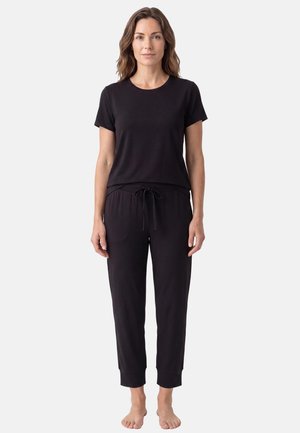 Femme debout pieds nus portant un t-shirt noir à manches courtes et un pantalon noir à cordon sur un fond clair uni.
