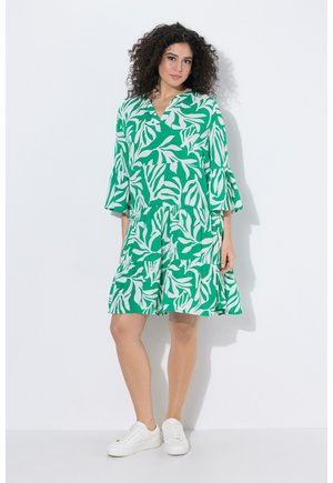 Frau mit lockigem Haar, die ein knielanges Kleid mit grün-weißem Blattmuster, ausgestellten Ärmeln und weißen Sneakers trägt, steht an einer weißen Wand.