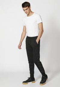 Sorte sporty joggers med en tapered fit og elastisk talje, parret med en tætsiddende hvid skjorte og sorte sneakers med gule accenter.