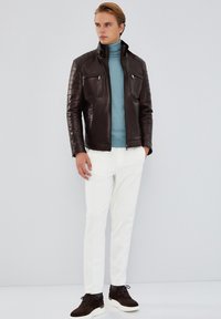 Giacca di pelle marrone con spalle trapuntate, tasche con zip; maglione azzurro chiaro, pantaloni bianchi e sneakers marroni scuri.