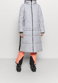 Personne portant un long manteau d'hiver matelassé gris avec des détails noirs et néon sur les côtés, un pantalon de ski corail et noir, et des chaussures de ski noires.