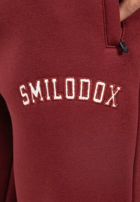 Bordeaux joggers van zacht materiaal, met witte geborduurde "SMILODOX" tekst, een ritszak en een geribbelde textuur.