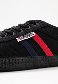 Black sneaker with black laces, featuring red and blue stripes on the side, a textured black sole, and a logo label near the laces.