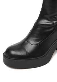 Botină din piele neagră până la gleznă, cu un design elegant, vârf ascuțit și o talpă groasă neagră, decorată cu detalii subtile de cusătură.