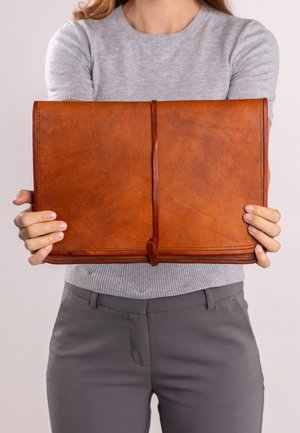 Braune Leder-Clutch mit glatter Oberfläche, rechteckiger Form und einer zentralen Verschlusslasche; gehalten von einer Person in einem grauen Pullover.