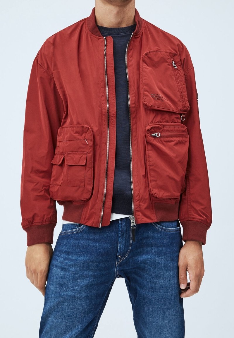 Homme portant une veste bomber rouge avec plusieurs grandes poches zippées et à rabat, un pull marine en dessous et un jean bleu.