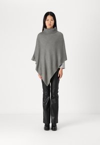 Grå stickad poncho med hög krage, med en slät textur och ett diamantmönster, bärs över svarta imitationläderbyxor.