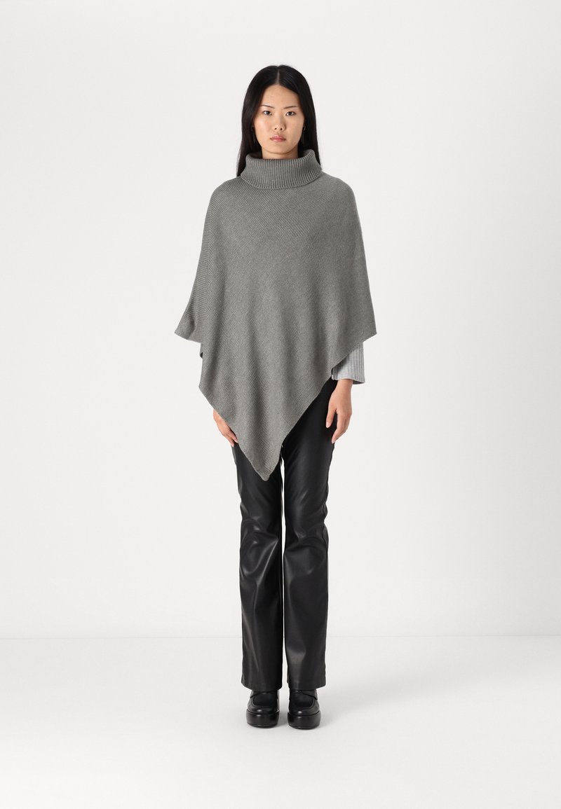 Grå stickad poncho med hög krage, med en slät textur och ett diamantmönster, bärs över svarta imitationläderbyxor.