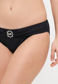 Bas de bikini noir avec un design froncé, arborant un logo MK argenté au centre devant. Texture lisse et forme taille moyenne.