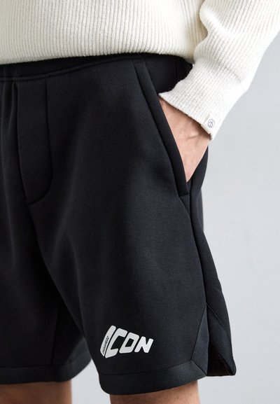 Shorts de sport noirs en tissu doux, dotés de poches latérales, d'un logo blanc sur la jambe et d'une aération discrète à l'ourlet.
