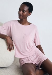 Kvinna med kort hår som bär en rosa kortärmad topp och rosa shorts med prickar, sittande och lutande mot en strukturerad vit kudde.