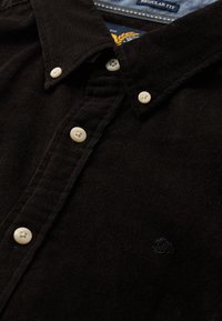 Camisa de pana negra con cuello abotonado, botones en color crema y sutiles rayas texturizadas. Pequeño logo bordado en el pecho.