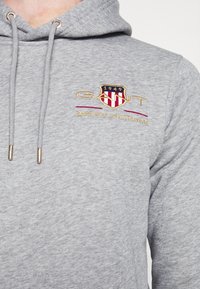 Grå hoodie med snore og metalender. Har broderet logo i rød, guld og marineblå. Blød tekstur og afslappet design.