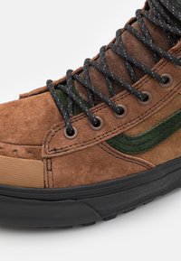 Bruna mockaskor med svarta snören, gröna mockadetaljer och en texturerad svart gummisula på vit bakgrund.