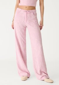 Femme portant un pantalon large brodé de fleurs roses avec taille à cordon et des chaussures à enfiler tissées beige sur fond blanc.