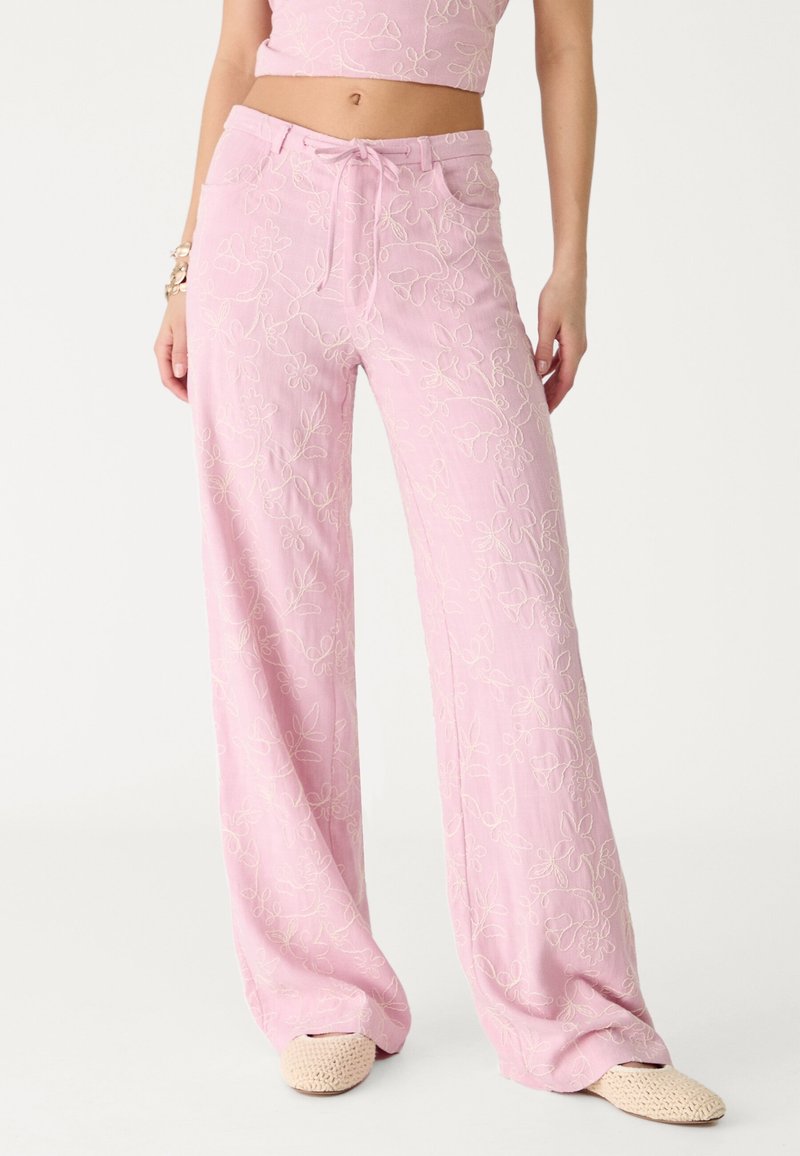 Femme portant un pantalon large brodé de fleurs roses avec taille à cordon et des chaussures à enfiler tissées beige sur fond blanc.