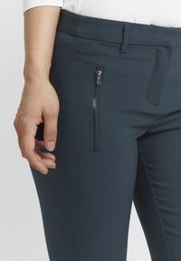 Námořnické modré kalhoty vyrobené z elastické tkaniny s úzkým střihem. Obsahují kapsu na zip a boční švy, což zajišťuje čistý a upravený vzhled.