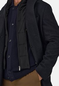 Dunkler Wollmantel über einer schwarzen gesteppte Weste, darunter ein dunkelblauer gerippter Pullover. Mit Knöpfen und einer glatten Textur.