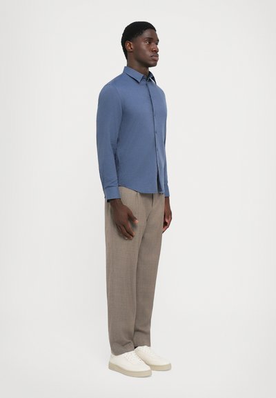 Man staat rechtop en draagt een lichtblauw overhemd met lange mouwen, beige broek en witte sneakers tegen een eenvoudige witte achtergrond.