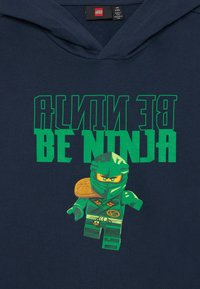 Sweatshirt azul-marinho com um gráfico de um ninja LEGO verde e o texto verde "SEJA NINJA". O design destaca a armadura e os acessórios do ninja.