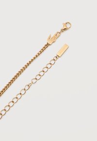 Χρυσή αλυσίδα με κούμπωμα σε σχήμα αστακού, μπρελόκ σε σχήμα αλιγάτορα και ορθογώνια ετικέτα με χαραγμένο το "LACOSTE".