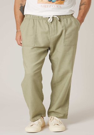 Homme portant un pantalon décontracté vert clair avec taille à cordon, poches appliquées, baskets blanches et t-shirt blanc avec motif Arizona.