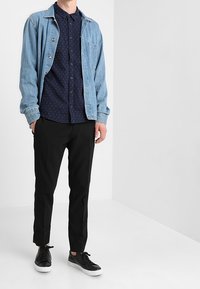 Denim jack over een marineblauwe knoopshirt met een klein patroon, gecombineerd met zwarte tappered broeken en zwarte sneakers met witte zolen.