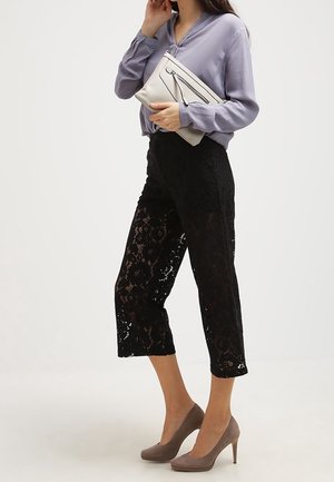 Mujer con pantalones cortos de encaje negros, blusa lavanda, tacones altos topo, sosteniendo un bolso de mano beige contra un fondo blanco.