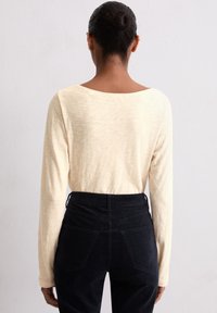 Chemise beige clair à manches longues avec un large décolleté, associée à un pantalon en velours côtelé noir taille haute avec poches arrière et texture subtile.
