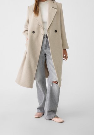 Kobieta ubrana w beżowy długi płaszcz, biały prążkowany sweter, podarte jasnoszare dżinsy oraz jasnoróżowe baleriny stojąca na jednolitym tle.