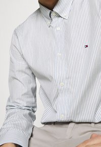 Světle modrá košile na knoflíky s tenkými vertikálními pruhy. Obsahuje malé logo na hrudi, bílé knoflíky a zaoblený límec. Hladká textura.