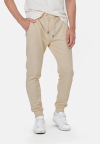 Beige Sweatpants aus elastischem Material, schmal zulaufend, mit elastischem Bund und Kordelzug sowie Seitentaschen, kombiniert mit weißen Sneakers.