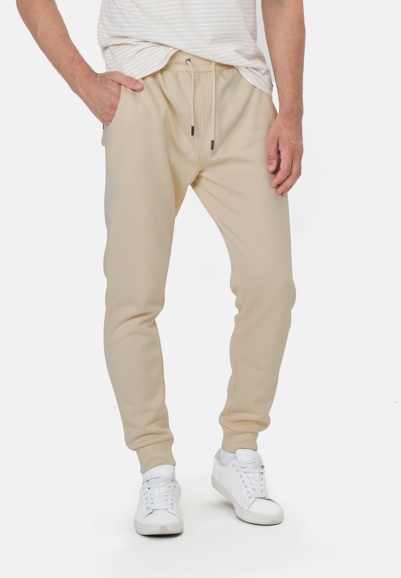 Beige Sweatpants aus elastischem Material, schmal zulaufend, mit elastischem Bund und Kordelzug sowie Seitentaschen, kombiniert mit weißen Sneakers.