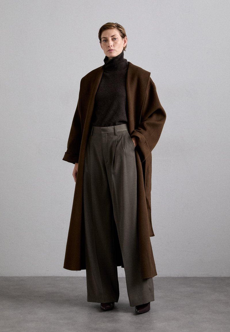 Cappotto oversize marrone sopra un dolcevita scuro, abbinato a pantaloni a gamba larga grigi a vita alta, caratterizzati da linee pulite e un tessuto texture.