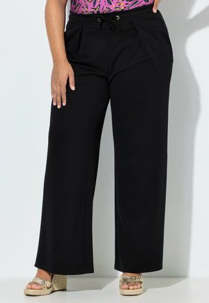 Frau trägt schwarze Hose mit weitem Bein und Kordelzug in der Taille, bunte gemusterte Bluse und goldene Keilsandalen, steht vor einer weißen Wand.