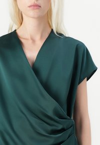 Vihreä satiiniwrap-toppi lyhyillä hihoilla, jossa on V-kaula-aukko ja pehmeät laskokset edessä. Sileä pinta ja hienovarainen kiilto.