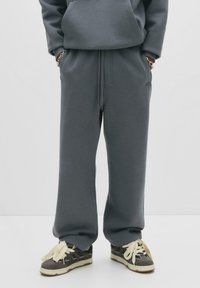 Person i løse grå sweatpants og mørke sneakers med tykke cremefarvede snørebånd, hænderne stukket i lommerne mod hvid baggrund.