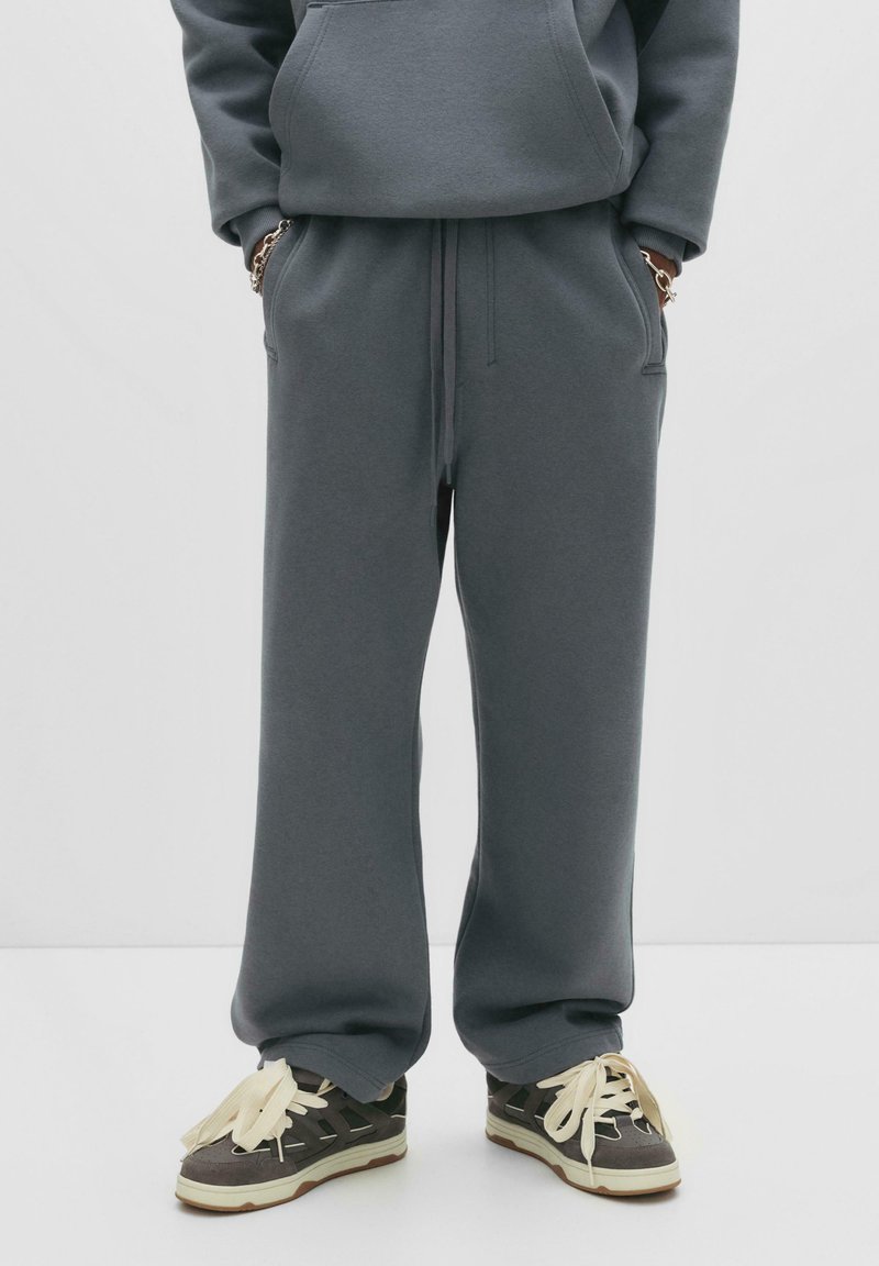 Person i løse grå sweatpants og mørke sneakers med tykke cremefarvede snørebånd, hænderne stukket i lommerne mod hvid baggrund.