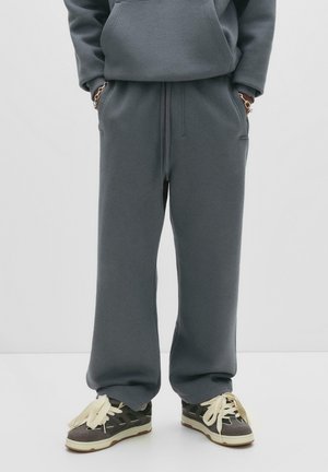 Személy laza szürke melegítőnadrágban és sötét sportcipőben vastag krémszínű fűzőkkel, kezei a zsebében egy fehér háttér előtt.