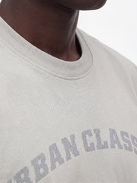 Zbliżenie osoby noszącej jasnoszarą koszulkę z częściowo widocznym wyblakłym tekstem "URBAN CLASS" na piersi.