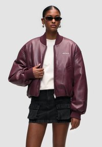 Burgundská kožená pilotní bunda s žebrovanými manžetami a límcem, s předním zipem a vyšívaným logem na hrudi, doplněná černou džínovou sukní.